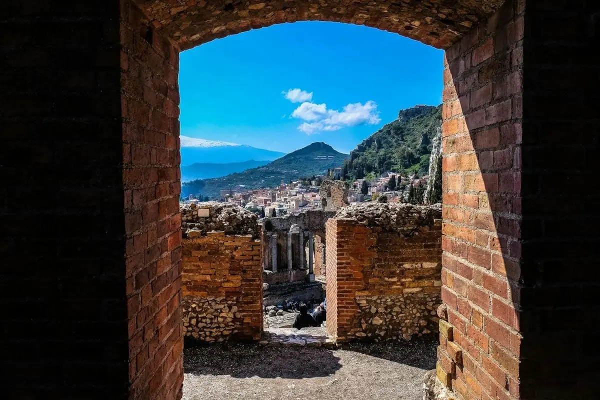 Old ruins viewed through a red brick arch, overlooking a scenic landscape and mountains under a blue sky.
