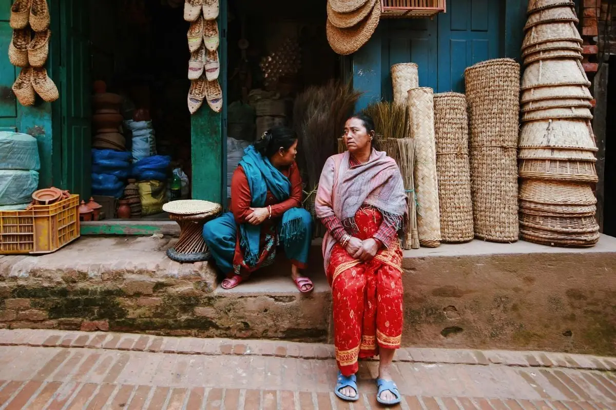 Two women in traditional attire sit outside a colorful shop selling baskets and woven goods in a village.