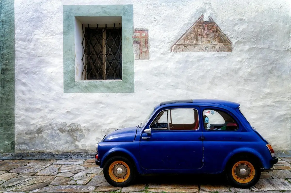 A classic blue car parked by a textured wall with historical architecture.