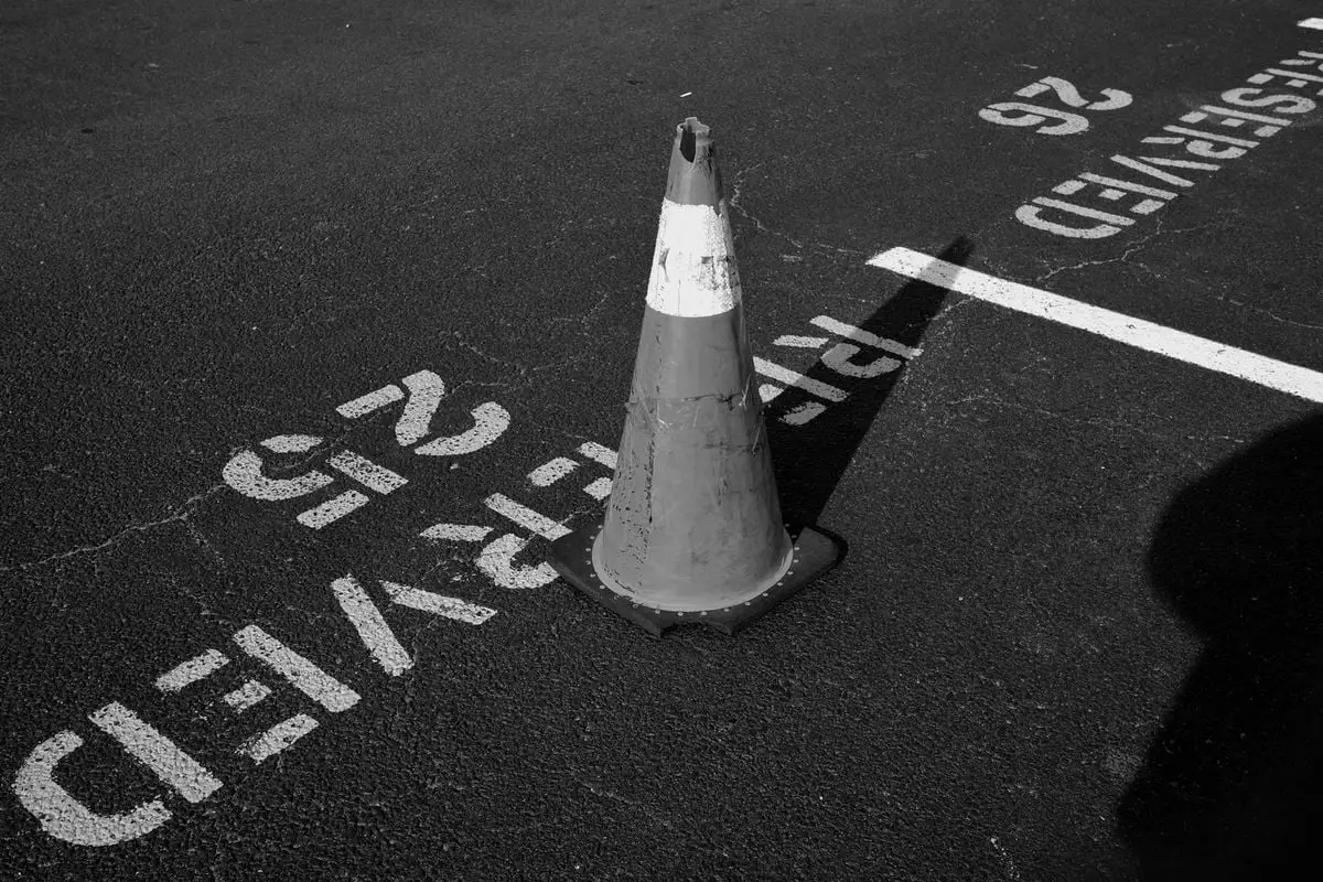 Black and white image of a single traffic cone on marked parking lot asphalt.