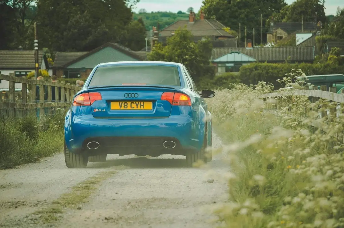 A blue Audi drives on a rural road through a small town with houses around.