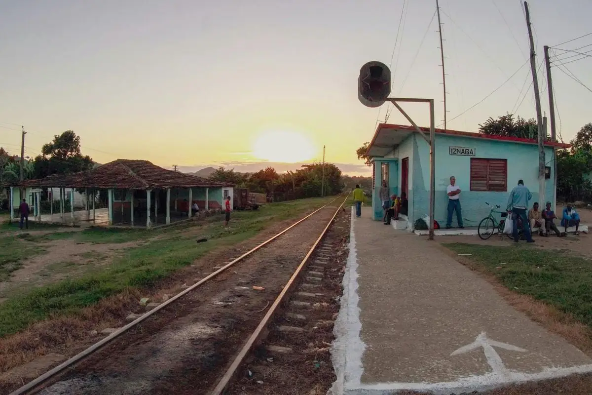 A rustic train station in the countryside during sunset, lively with locals gathering around.