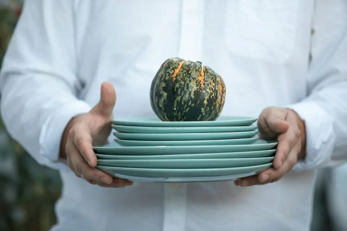 Person holding a stack of green plates with a small squash on top, symbolizing fall.