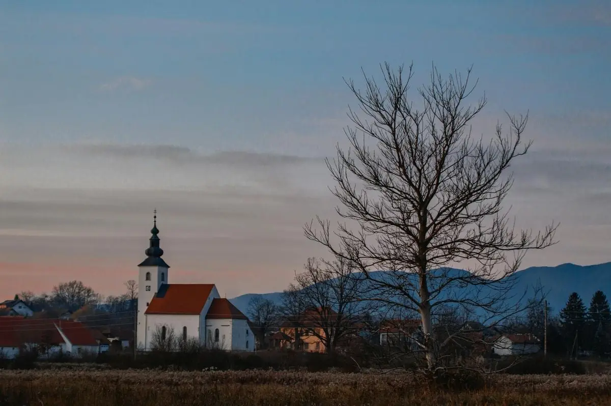 Charming village scene with a church and tree set against a mountain backdrop at sunset.