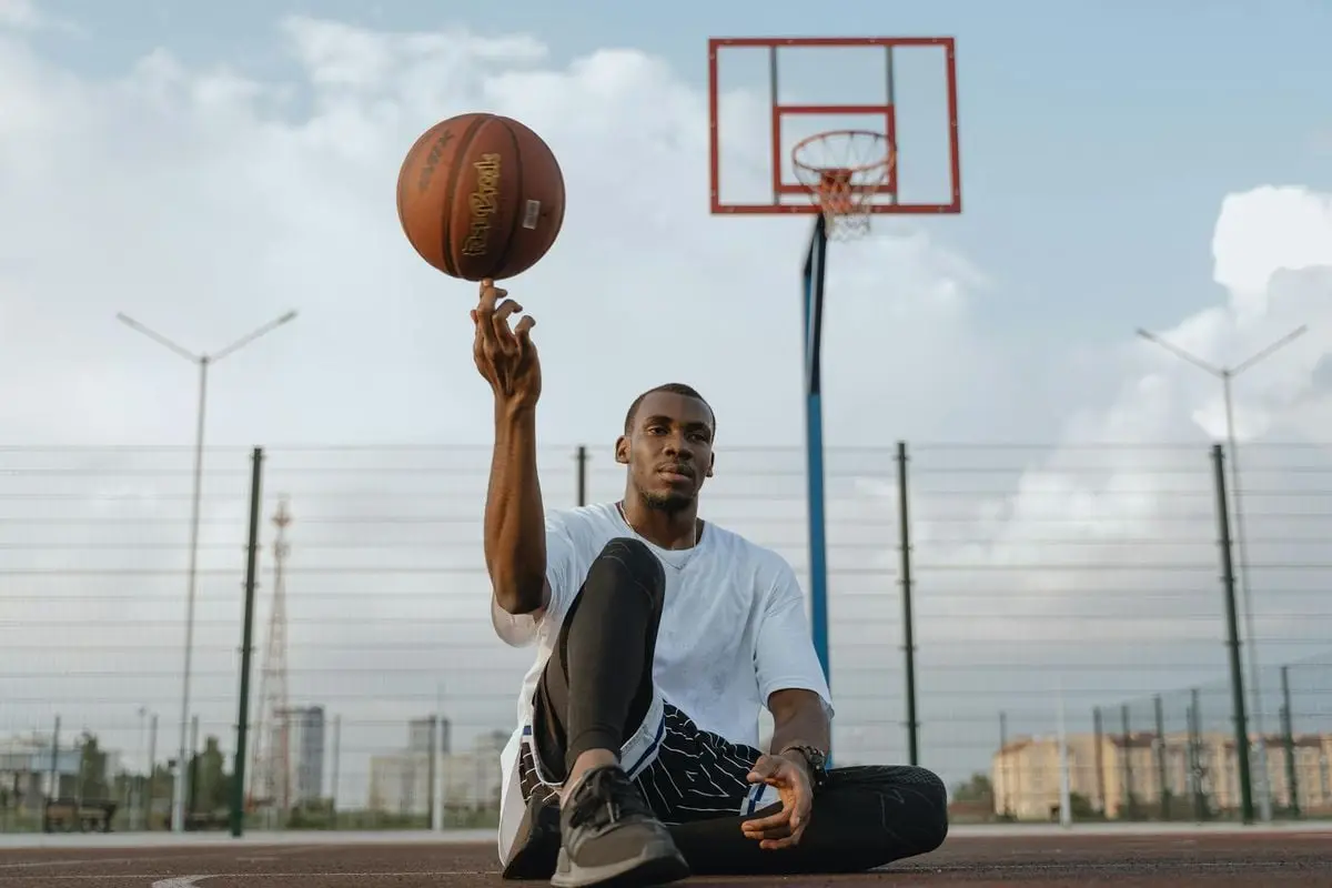 A young black man balancing a basketball on his finger while sitting outdoors on a court.