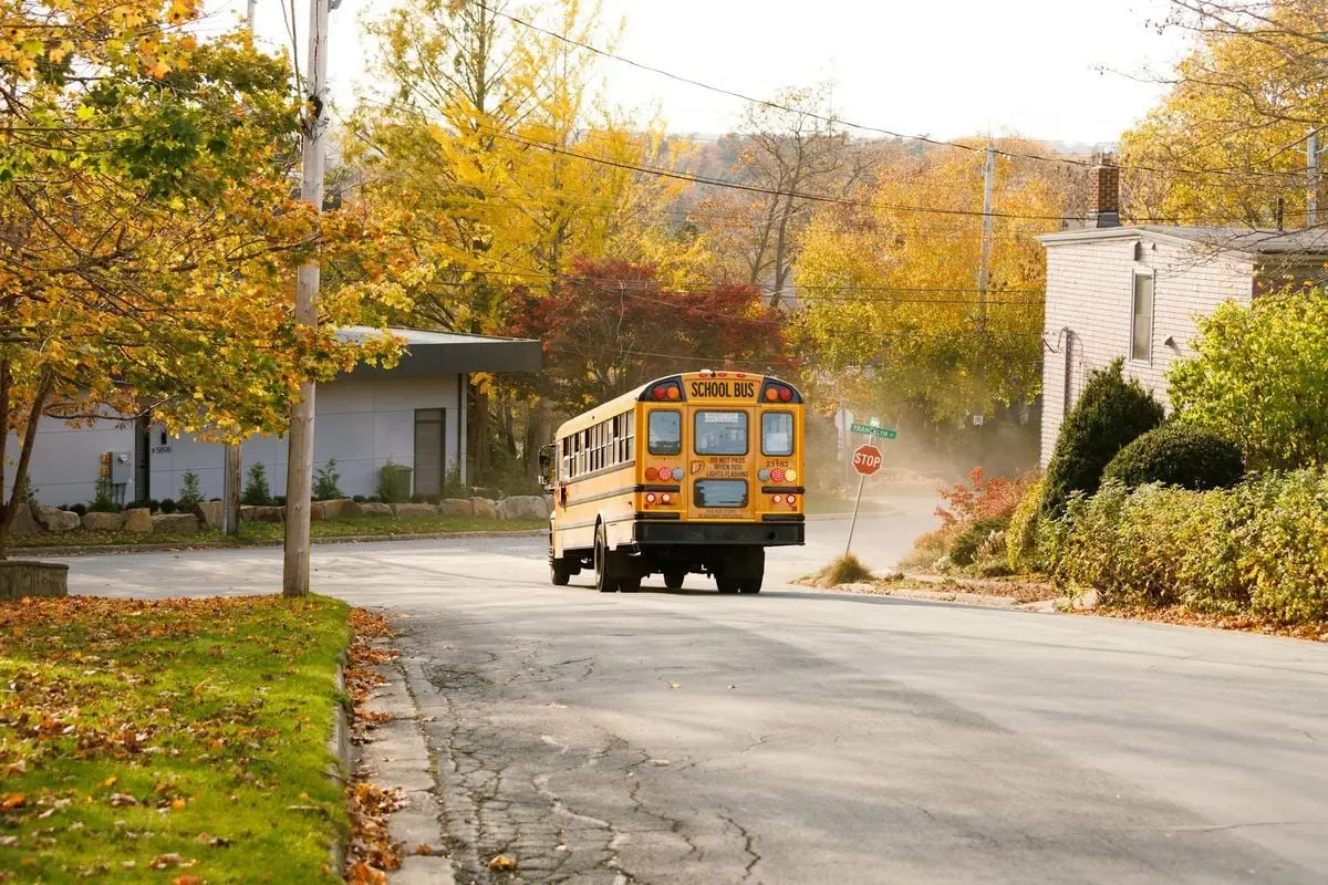 A yellow school bus on a village road during a crisp autumn day, surrounded by colorful trees.