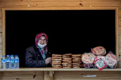 An elderly woman selling traditional bread and water from a wooden stall in an outdoor street market.