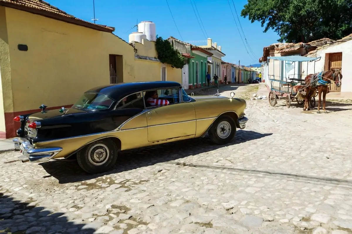 Vintage car passing by colorful houses and horse cart on a sunny cobblestone street.