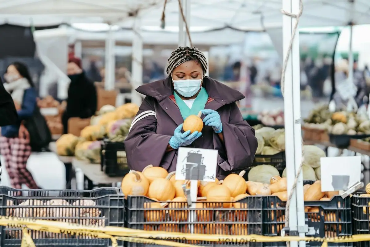Adult African American female nurse in medical uniform and gloves with mask choosing pumpkin from box while standing in daylight in street market