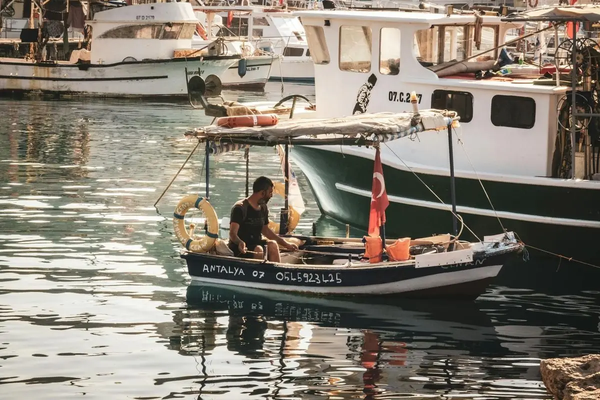 A man sits on a small motorboat with a Turkish flag in Antalya harbor, surrounded by other vessels.