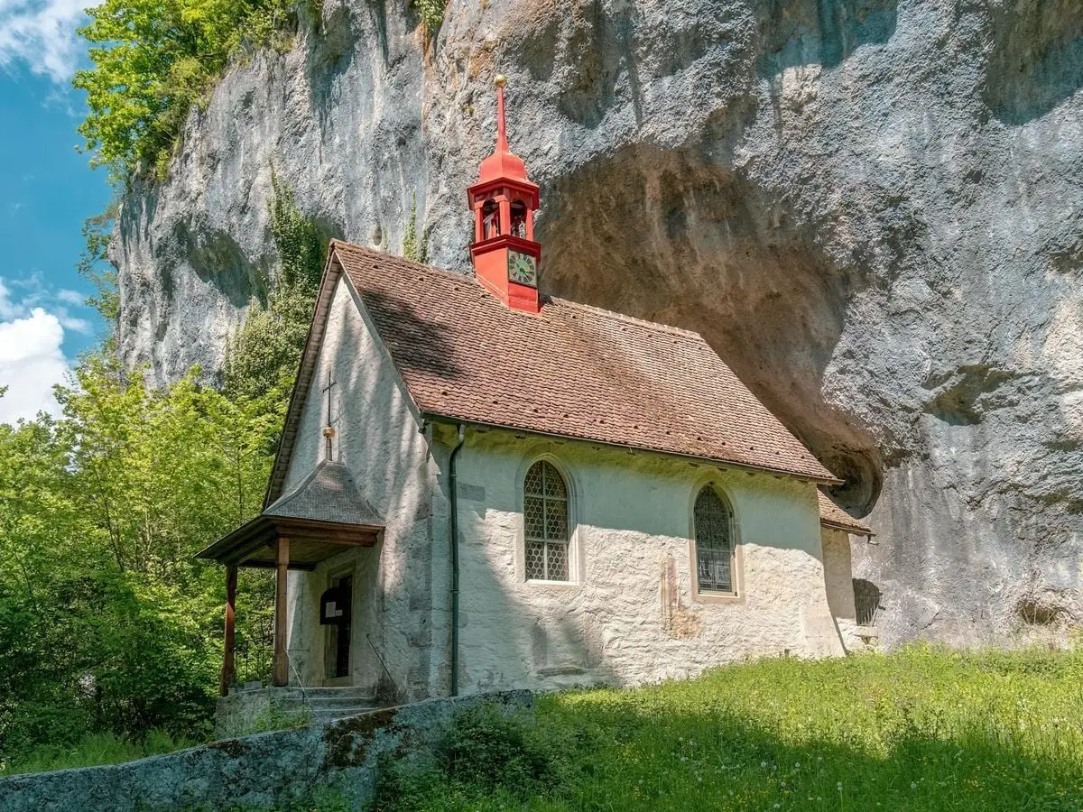 Explore a picturesque historic chapel nestled against a cliff, surrounded by lush greenery under a bright sky.