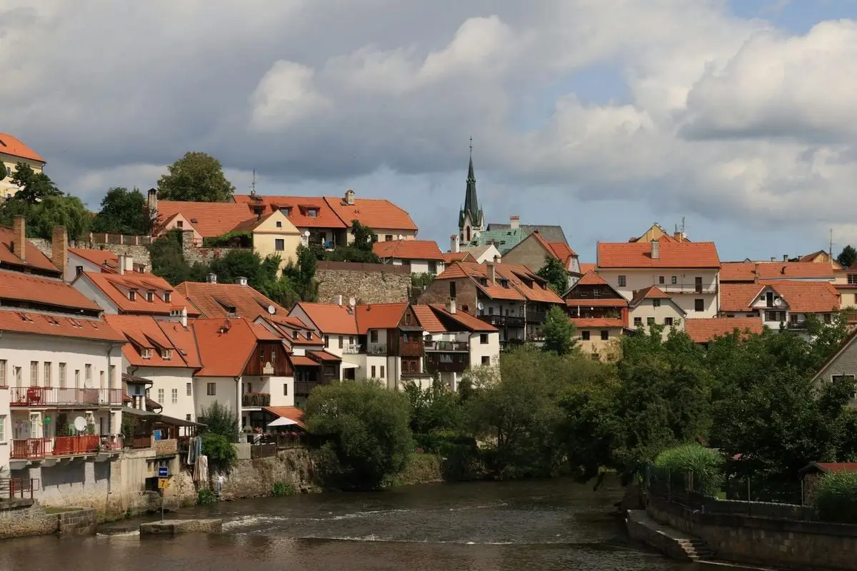 Picturesque European village with red-tiled roofs along a serene river under a partly cloudy sky.