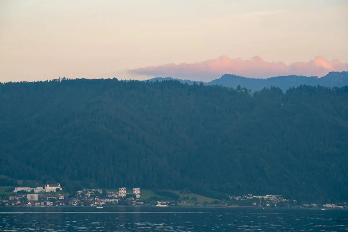 Peaceful lakeside town with lush forested mountains against pastel skies.