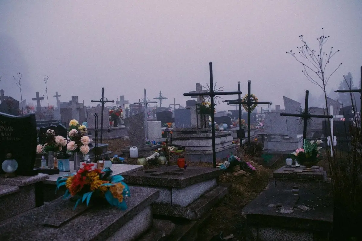 A fog-covered cemetery at sunrise with gravestones, crosses, and flower arrangements, evoking a somber mood.