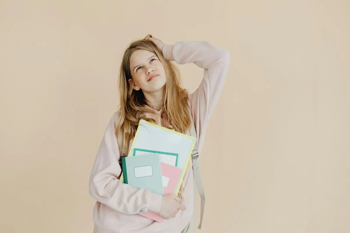 Teenage girl in pink hoodie holding colorful notebooks against beige background.