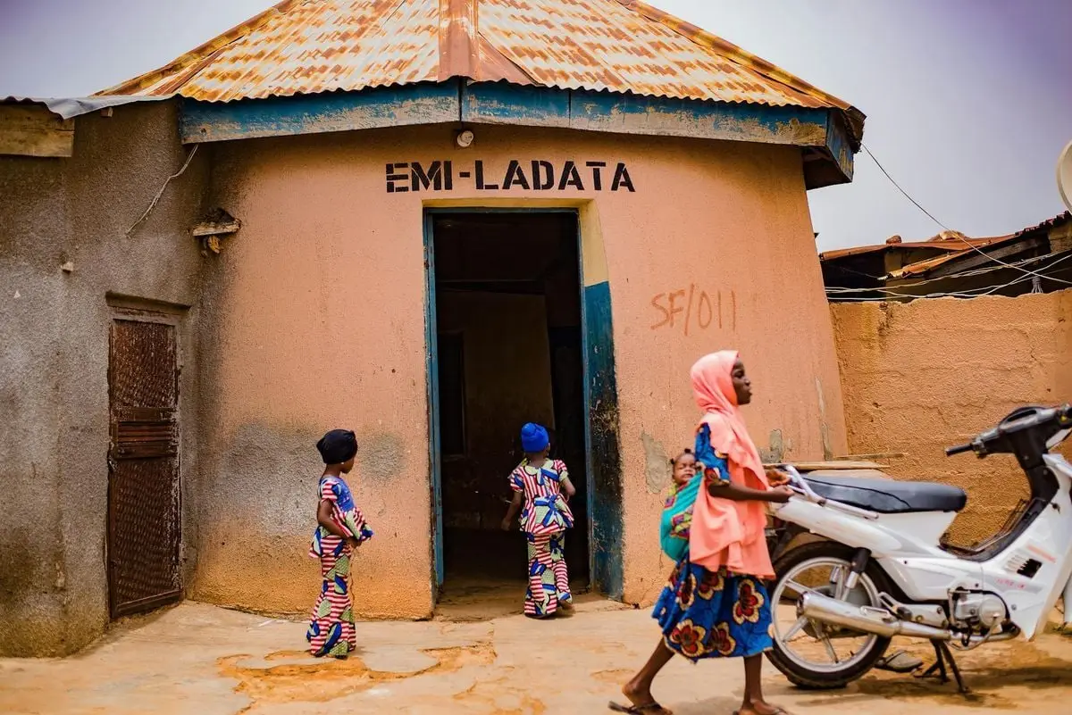 Children playing outside a rustic building with a woman passing by.