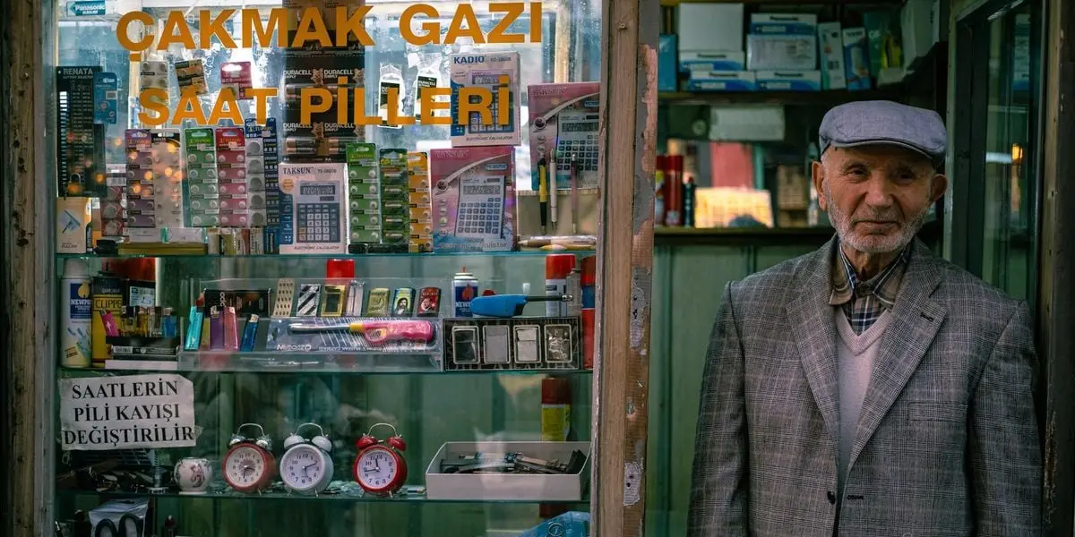 Elderly man stands by a vintage electronics shop window displaying various batteries and gadgets.