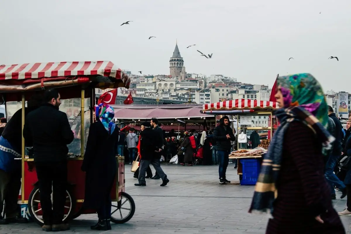 Vibrant street market scene in Istanbul with crowds, kiosks, and the iconic Galata Tower in the background.