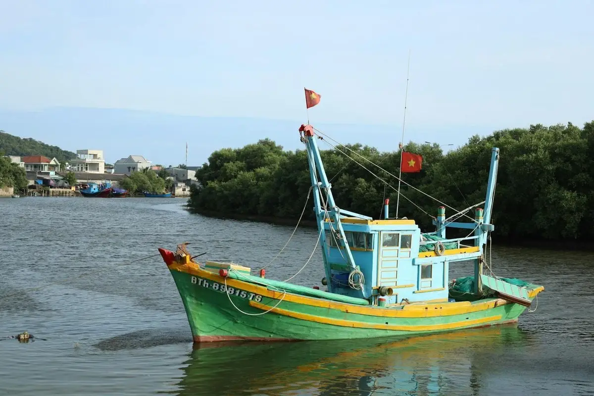 A vibrant fishing boat floating on a calm river, with lush greenery in the background.
