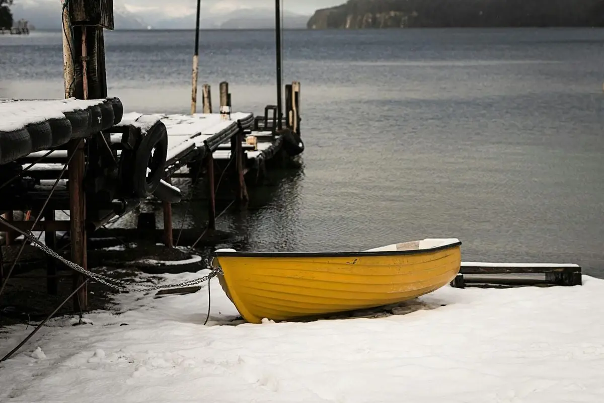 A lone yellow rowboat rests on snow by a wooden dock at a serene lake in Villa La Angostura, Argentina.