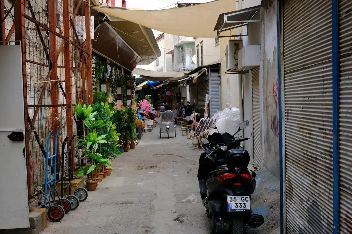 Explore a quaint alley in İzmir with a parked motor scooter, local shops, and vibrant plants.