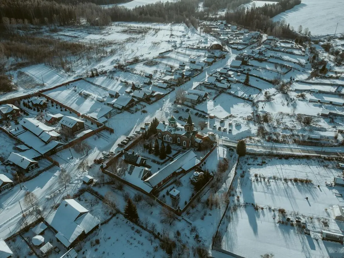 Aerial shot of a snow-covered rural village with scattered houses and a church surrounded by snowy fields.