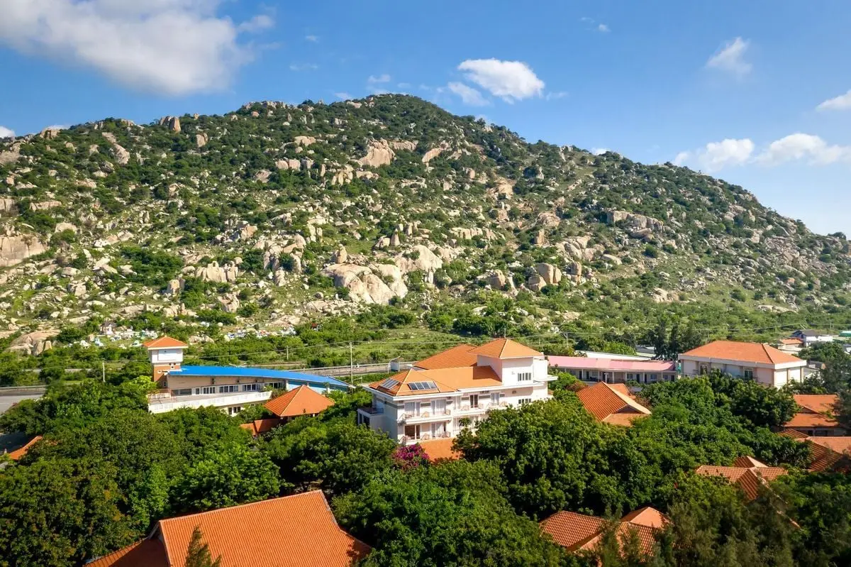 Aerial view of a sunny town with red-tiled roofs at the base of a green mountain.