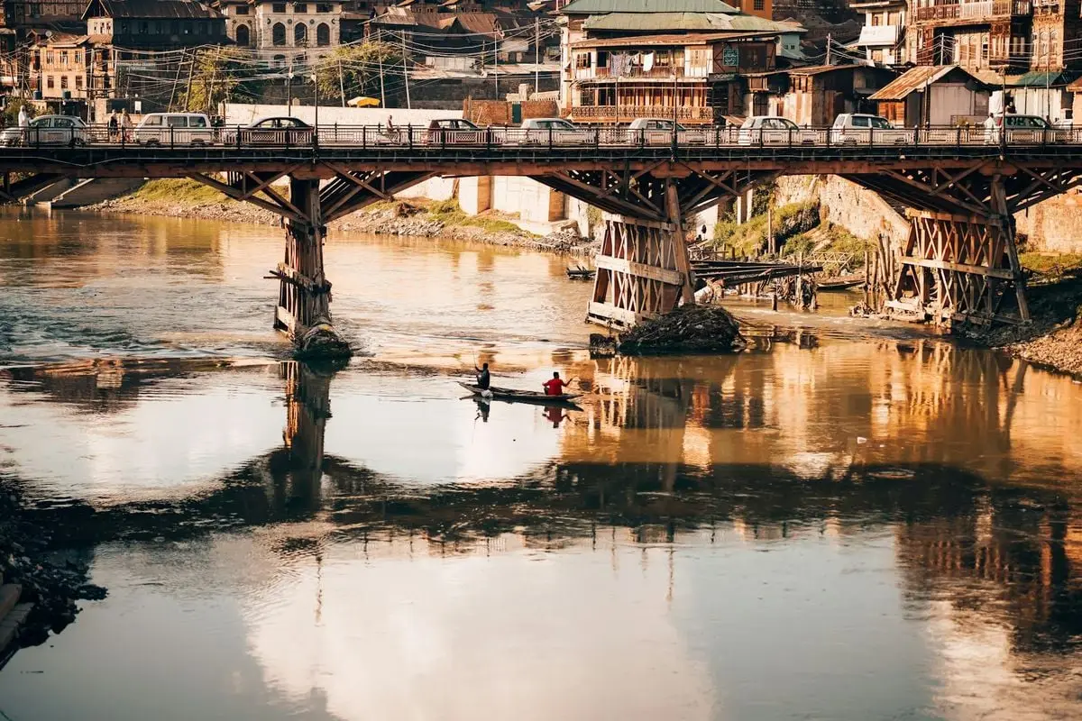 A canoe with two people under a wooden bridge spans a tranquil river in a rustic urban setting.