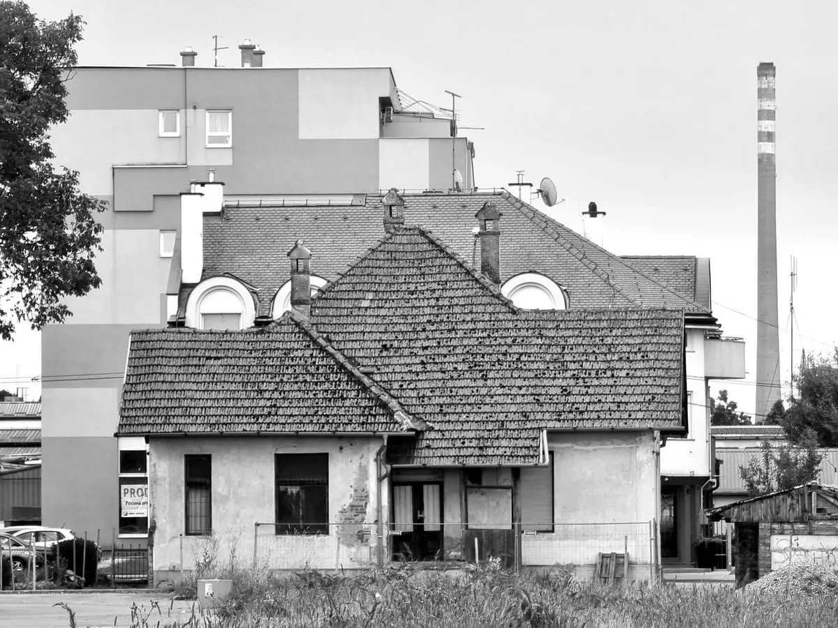 Black and white photo of a rustic house in Kutina, Croatia, framed by urban buildings and a chimney.