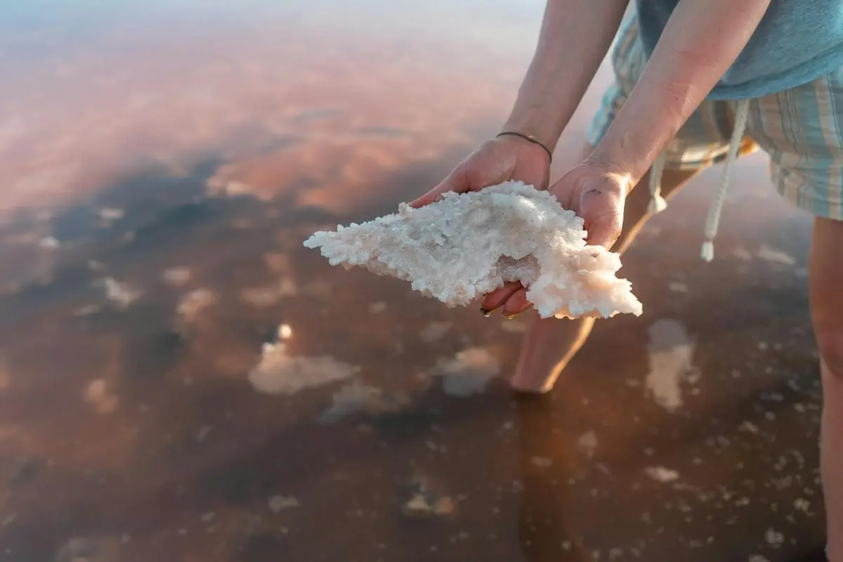 Close-up of hands holding salt crystals from a shallow saline lake at sunset.