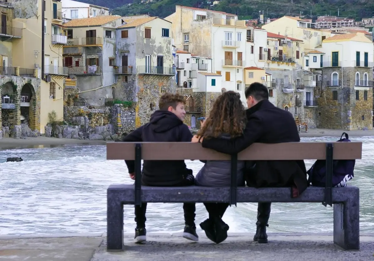 Three friends relax on a bench, enjoying the view of Cefalù's picturesque coastal buildings in Sicily.