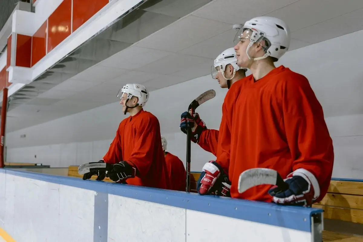 Three ice hockey players in red jerseys and helmets stand on an indoor rink bench.
