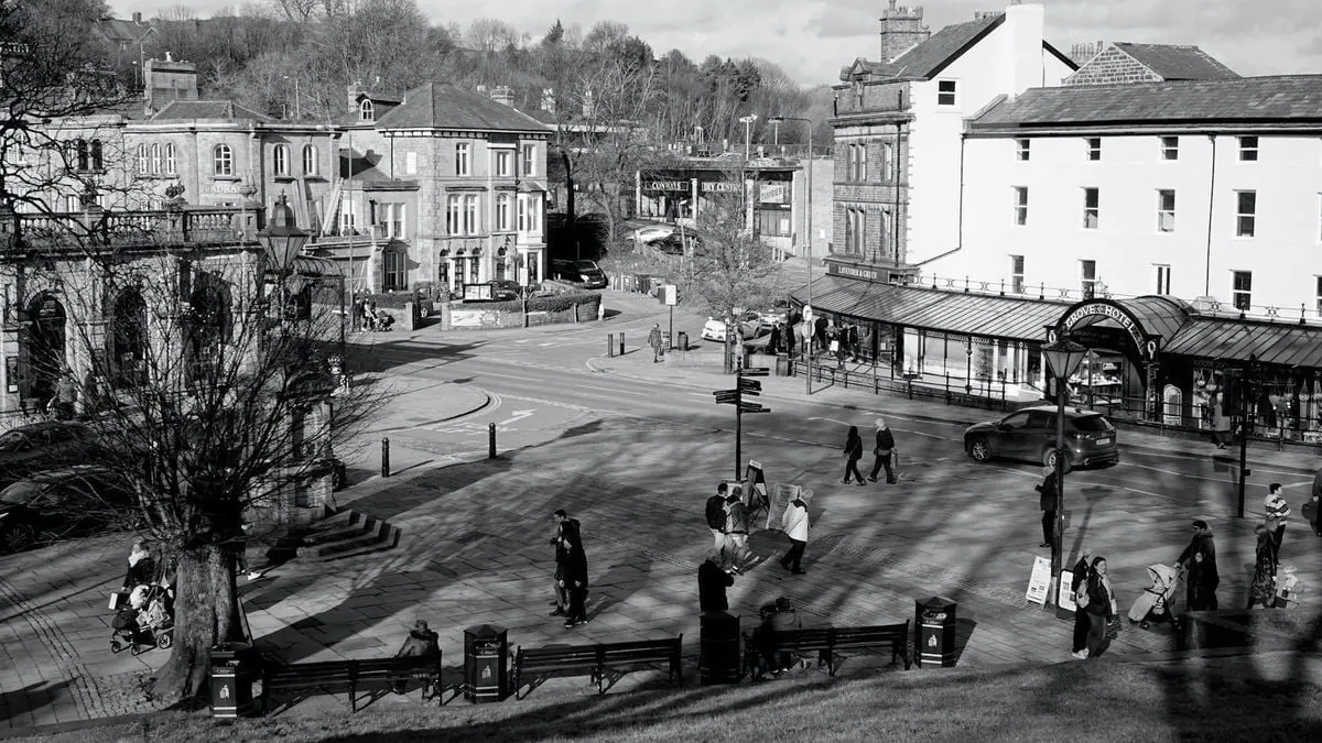 Black and white image of a busy street corner in Buxton, England, showcasing historical buildings and passersby.