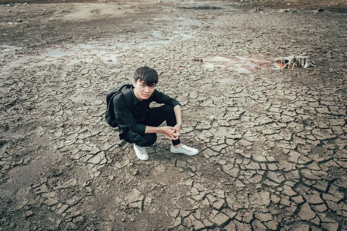 A young man wearing a backpack sits on cracked dry ground showcasing drought.