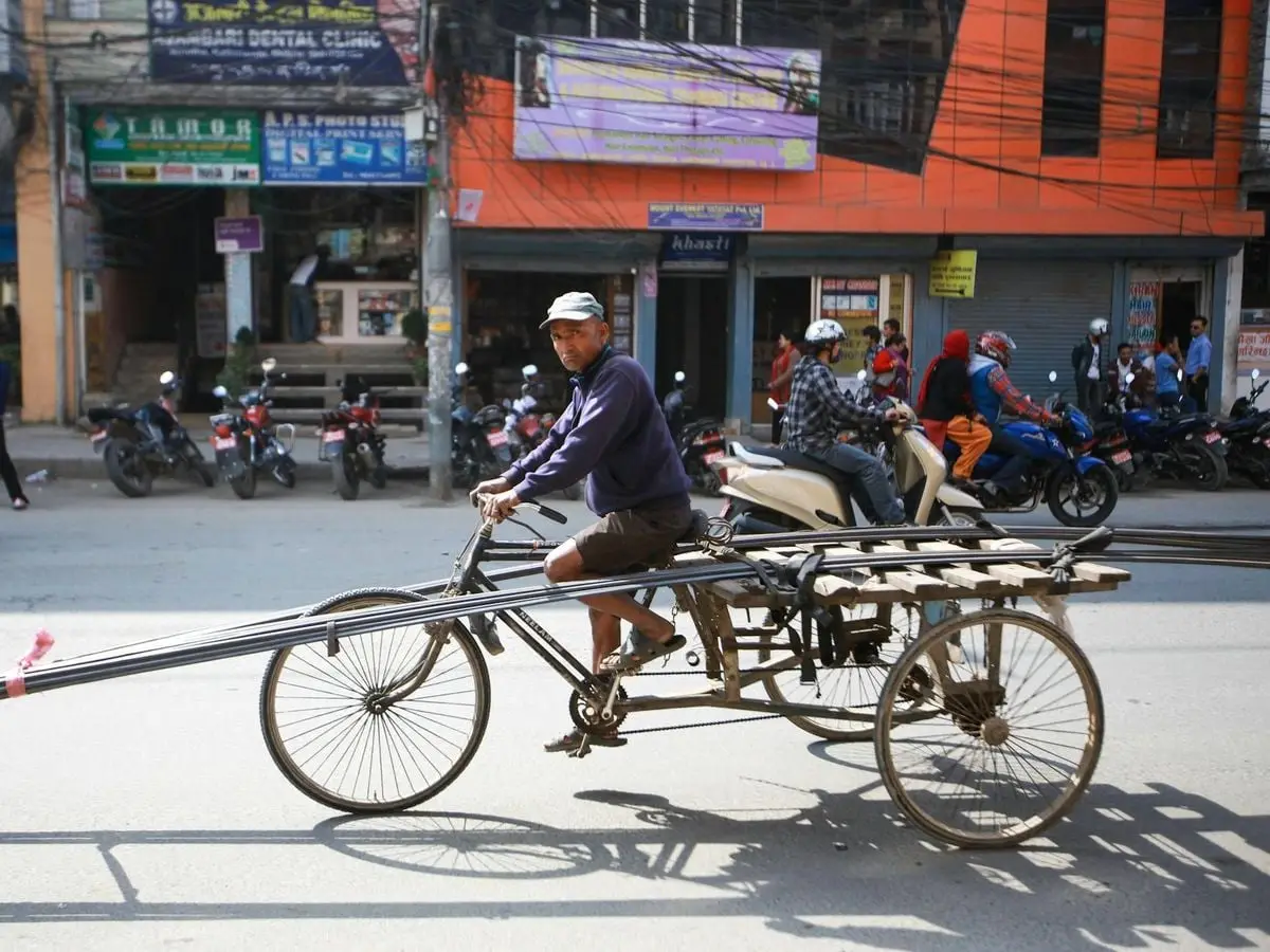 A man on a tricycle transporting metal rebars amidst city traffic, showcasing urban life.