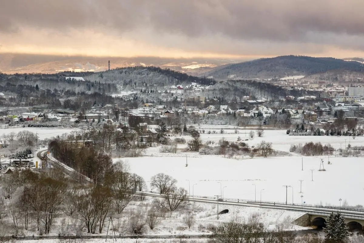 A serene winter landscape featuring a snow-covered town, distant mountains, and a bridge at dusk.