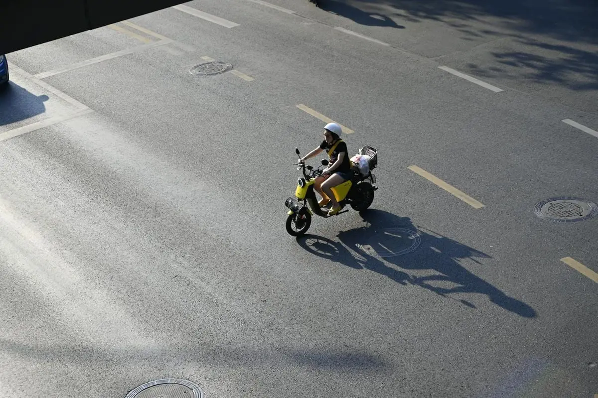 A woman riding a yellow scooter down a city street under bright daylight.