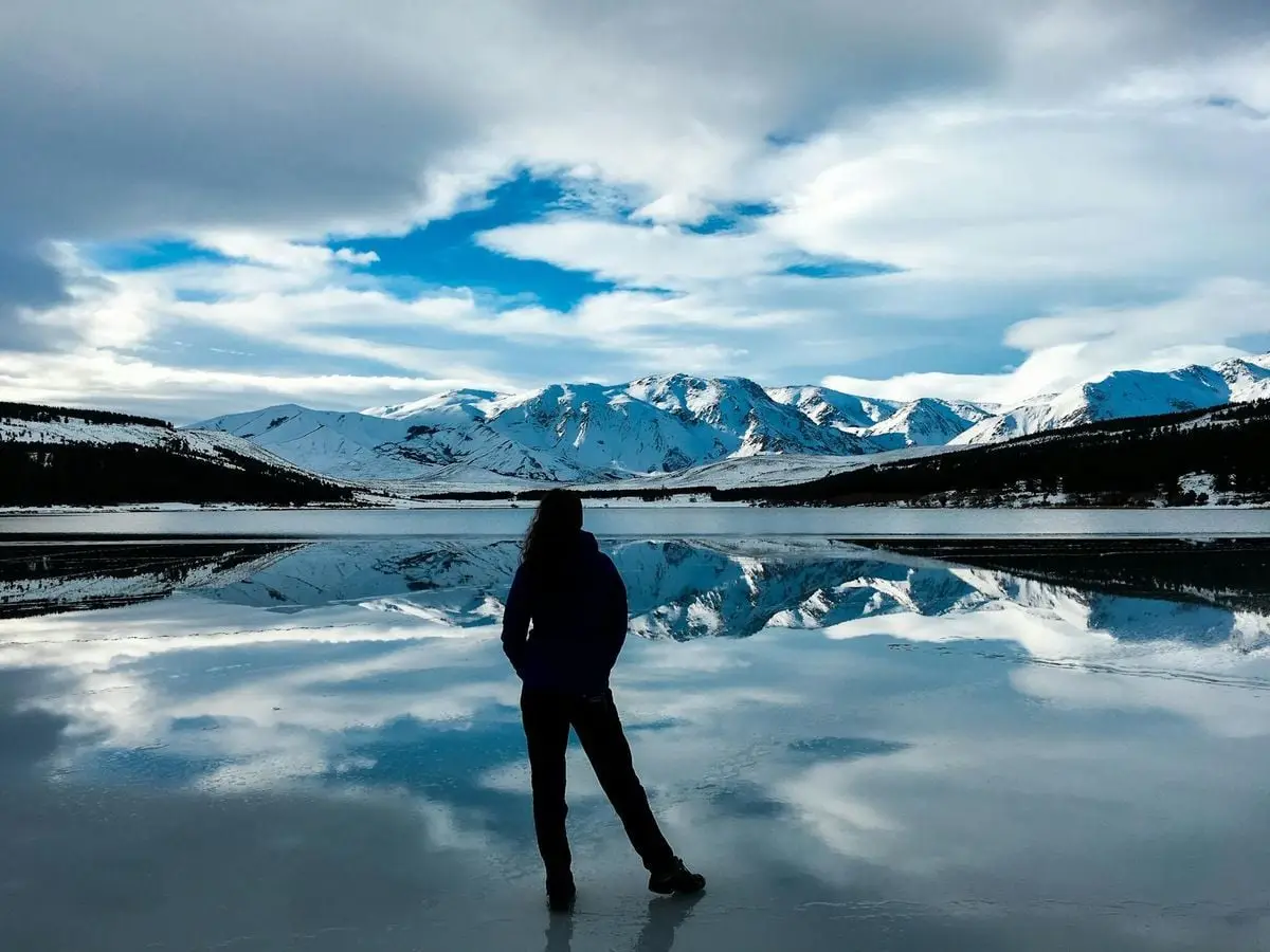 Back view of a woman standing on a frozen lake with reflections of snow-capped mountains in Patagonia, Argentina.
