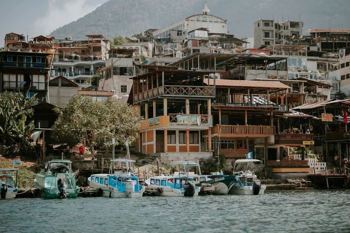 Lakeside view of a quaint town with colorful boats docked in front, nestled in a mountainous backdrop.