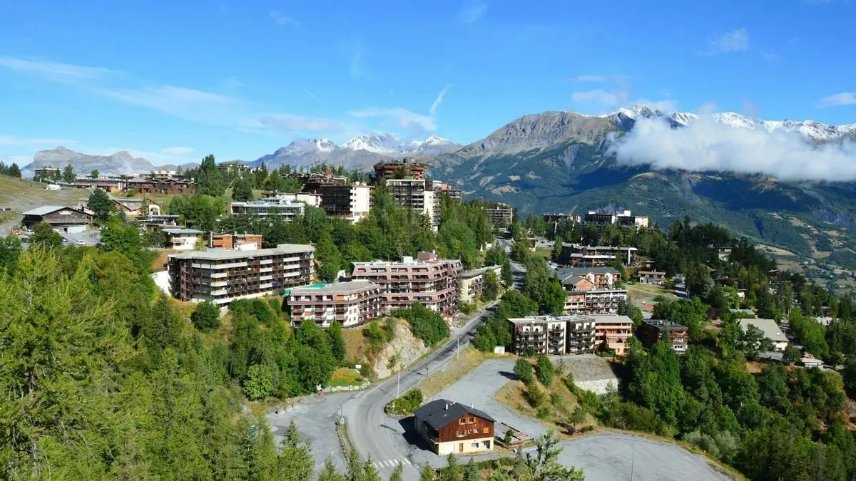 A picturesque alpine town with mountains, trees, and a clear blue sky is captured from above.