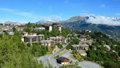 A picturesque alpine town with mountains, trees, and a clear blue sky is captured from above.