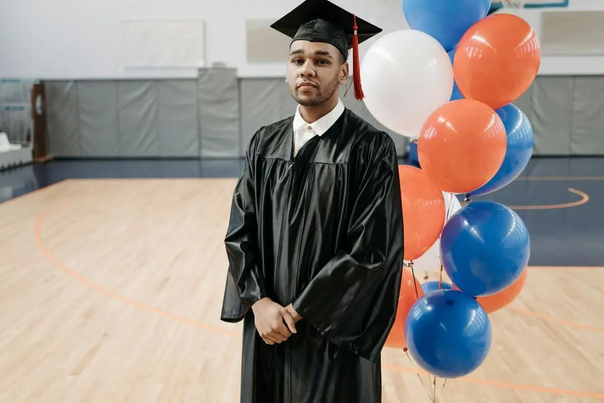 Young graduate in cap and gown posing in a gym with colorful balloons symbolizing achievement.