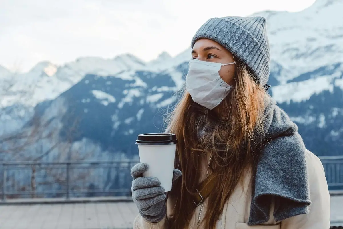 Woman in winter attire with coffee cup and face mask in snowy mountains.