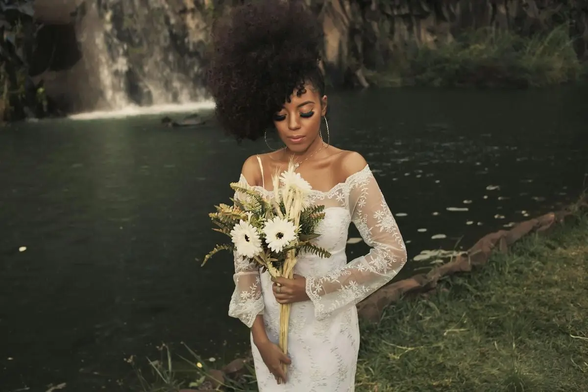 Beautiful bride in an elegant lace gown holding a bouquet by a waterfall, showcasing her unique hairstyle.