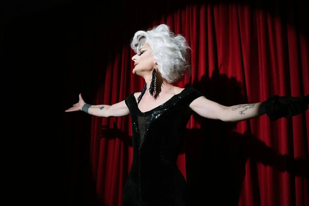 Stunning drag queen in a black dress performing on stage with a vibrant red curtain backdrop.