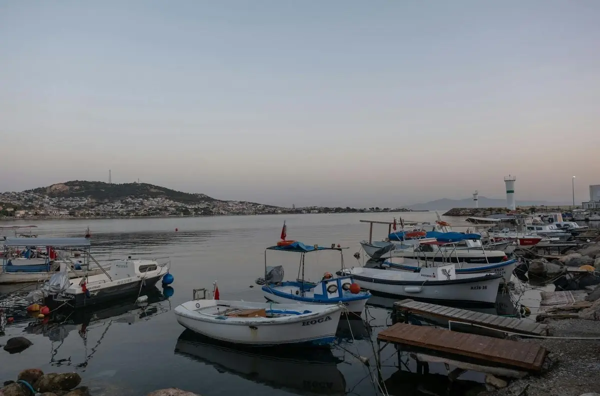 Serene view of fishing boats at Foça harbor during sunset with a distant lighthouse.