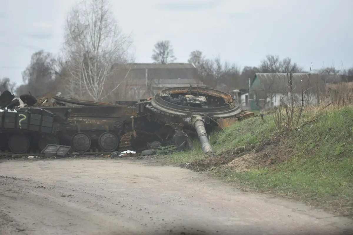 A destroyed military tank wreck on a dirt road near rural houses.