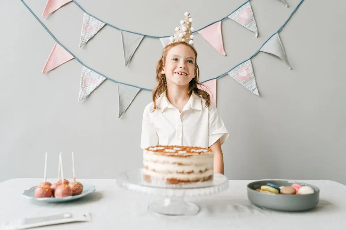A smiling girl at a birthday party with a cake, wearing a party hat indoors.