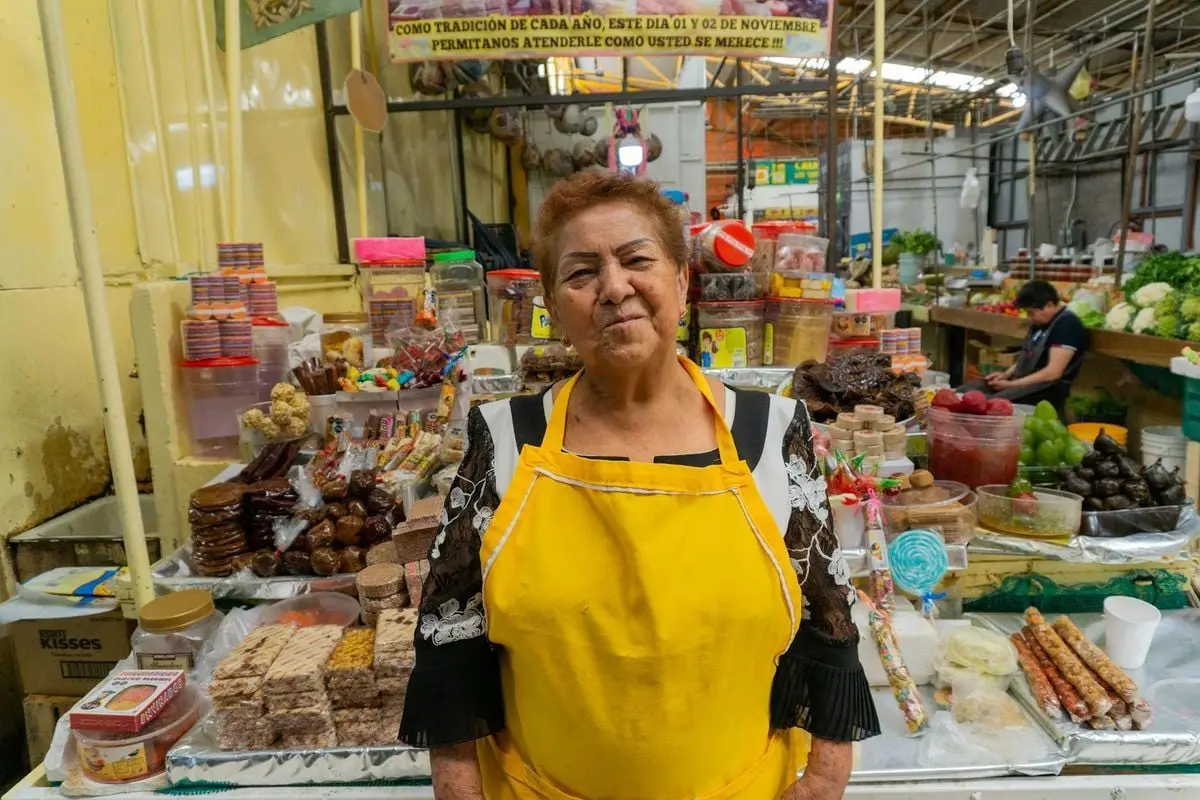 A cheerful woman in a yellow apron stands at her colorful market stall filled with local goods.