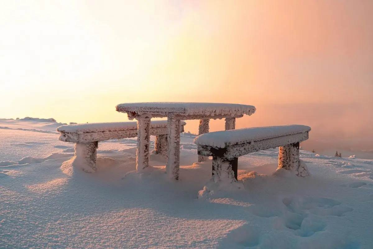 Snow-covered bench and table in a frosty winter sunset landscape.